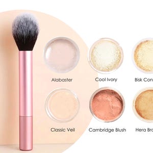 Op de afbeelding: Een roze make-up kwast met een zwart-witte borstelkop wordt getoond naast zes kleine potjes make-up. De potjes bevatten make-up tinten met de labels Alabaster, Cool Ivory, Bisk Concealer, Classic Veil, Cambridge Blush en Hera Bronzer.