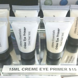 Puede incluir: Cinco tubos blancos de Ultimo Essentials Creme Eye Primer, cada uno con una tapa negra y una etiqueta que dice "Ultimo Essentials Creme Eye Primer 15ml Net Vol."