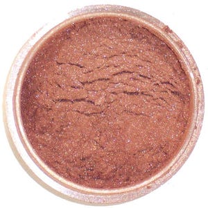 Può includere: Un vasetto di ombretto shimmer color bronzo. La polvere ha una superficie strutturata con brillantini visibili. Il contenitore è trasparente con un bordo colorato chiaro, che mostra il colore e la consistenza del prodotto.