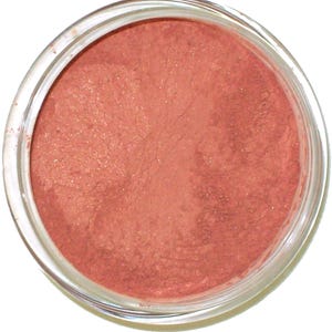 Può includere: Primo piano di un barattolo di vetro trasparente riempito con una polvere finemente macinata, scintillante, color pesca. La polvere ha un aspetto morbido e opaco con sottili particelle di glitter, che suggeriscono un prodotto cosmetico come blush o ombretto.