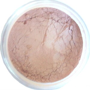 May include: A container filled with a light brown powder. The powder has a fine texture with visible cracks throughout. The container is clear, allowing the powder's color to be seen. The product appears to be a cosmetic item.