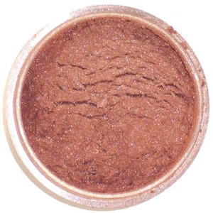 Può includere: Un piccolo contenitore rotondo pieno di polvere scintillante color bronzo. La polvere ha una superficie strutturata con sottili variazioni di colore e brillantezza. Il contenitore ha un bordo bianco e una base trasparente.