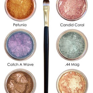 May include: Six small jars of shimmery eye shadow with names written below each jar: Petunia, Candid Coral, Catch A Wave, .44 Mag, Cleopatra, and Haut Apricot. A black makeup brush is in the center of the image.