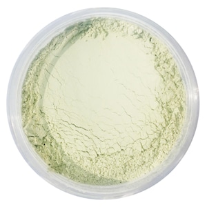 May include: A clear, round container filled with a pale green powder. The powder has a fine texture with visible cracks and imperfections. The container is transparent, allowing the powder's color to be fully visible. The product appears to be a cosmetic or skincare item.