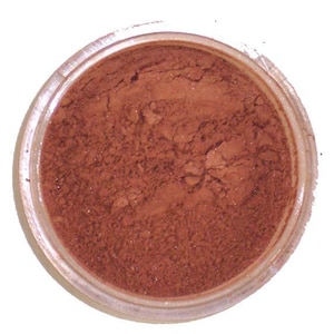 Può includere: Un piccolo barattolo trasparente pieno di polvere marrone-rossastra. La polvere ha un aspetto fine e strutturato. Il barattolo è posto su uno sfondo bianco. Il prodotto sembra essere un cosmetico, forse ombretto o fard.