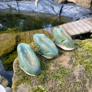 Puede incluir: Tres figuras de ranas de cerámica verdes, cada una con un diseño dorado único. Las ranas están colocadas sobre una roca cubierta de musgo, con un estanque y una pasarela de madera al fondo. Las ranas tienen la parte inferior blanca.