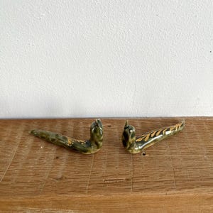 May include: Two ceramic, green and gold leaf-shaped decorative pieces. The pieces are curved and have a glossy finish. They are placed on a wooden surface.