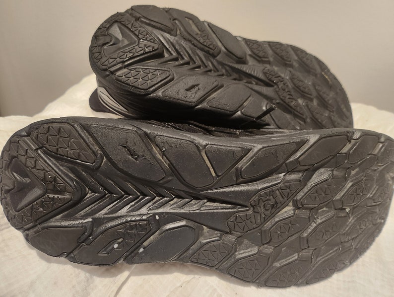 Puede incluir: Primer plano de las suelas de goma negra de un par de zapatillas para correr. Las suelas tienen un patr&oacute;n texturizado para el agarre.