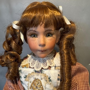 May include: A porcelain doll with auburn hair styled in pigtails, adorned with white ribbons. The doll has green eyes, freckles, and is wearing a dress with a gingham pattern and an apron with a beehive design and the text "Busy as a Bee".