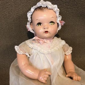 Puede incluir: Muñeca de bebé vintage con cara pintada, ojos verdes y un gorro de encaje blanco. La muñeca viste un vestido blanco con un lazo rosa y un cuello de encaje. Los brazos de la muñeca están extendidos.