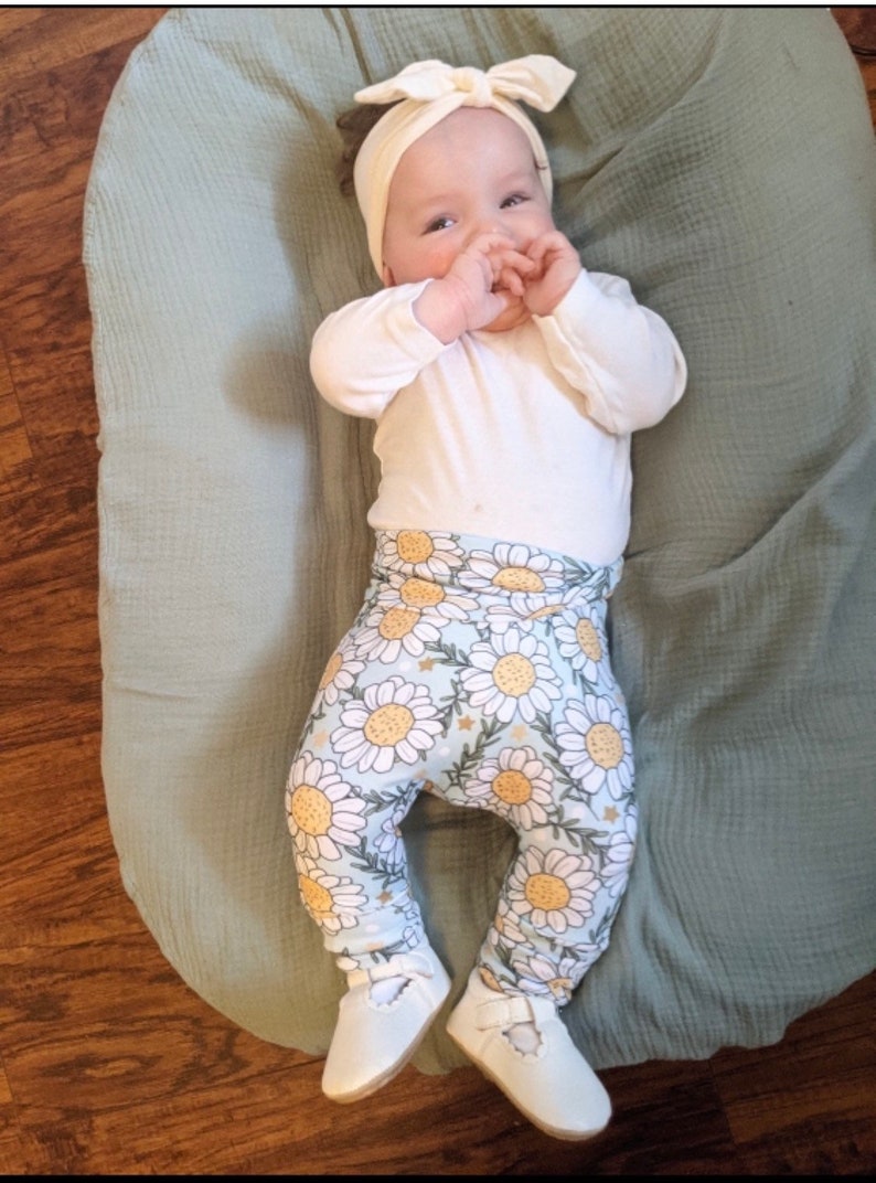 May include: A baby wearing a white long-sleeved shirt, white headband with a bow, and blue and white floral leggings. The baby is lying on a light green blanket.