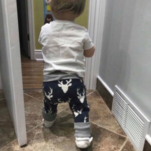May include: A toddler wearing a white shirt and blue and white patterned trousers with a deer design. The trousers have grey cuffs. The toddler is standing in a bathroom with white tiled floors.