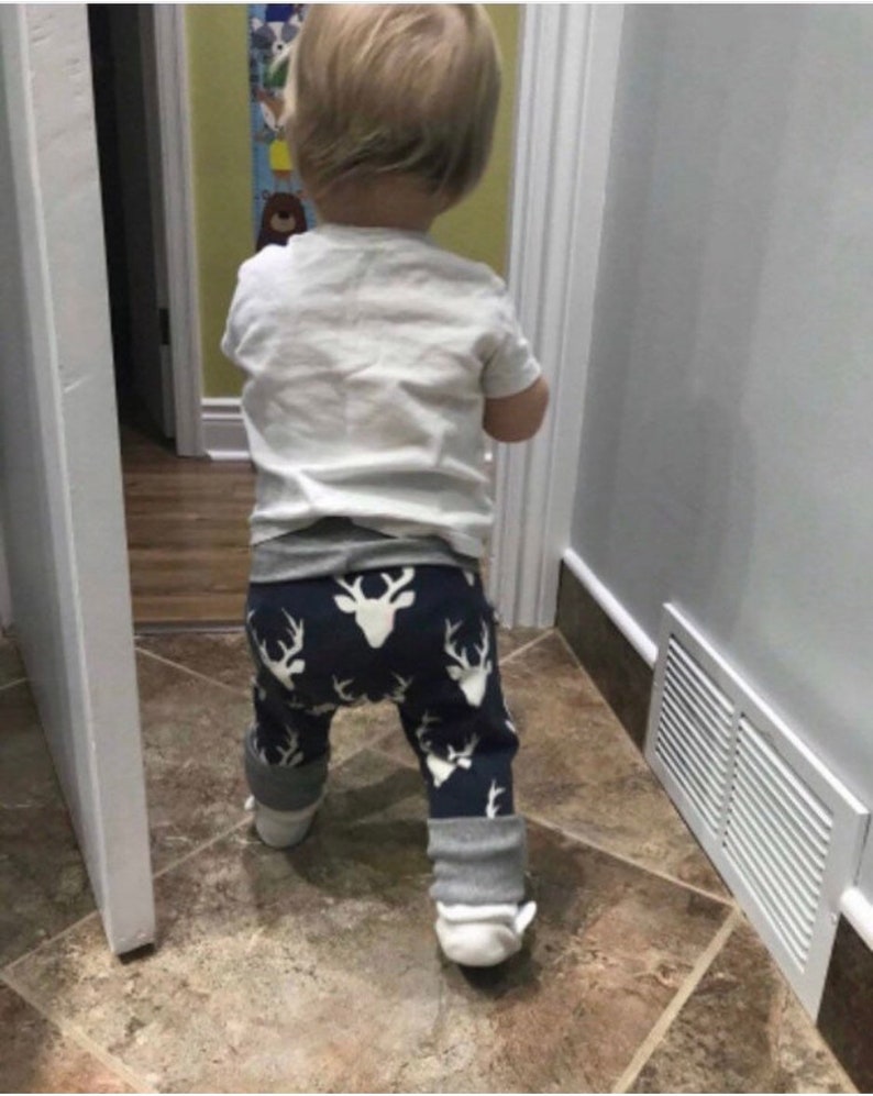 May include: A toddler wearing a white shirt and blue and white patterned trousers with a deer design. The trousers have grey cuffs. The toddler is standing in a bathroom with white tiled floors.
