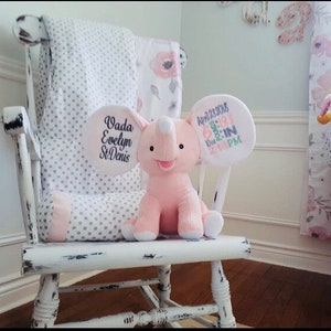 May include: A white rocking chair with a pink and grey polka dot blanket. A pink elephant plush toy is sitting on the blanket. The elephant has a white tag on its ear that says "Vada Evelyn St. Denis" and another tag that says "April 23, 2018 6:19 AM 7 lbs 12 oz 21" PM".