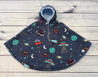 May include: A navy blue hooded poncho featuring a space-themed design with planets, stars, UFOs, and moons. The interior is light blue, and it has a zip closure. The poncho is designed for children.