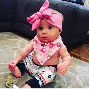 May include: A baby wearing a pink headband with a bow, a pink and grey elephant print bib, and pink and blue patterned leggings.