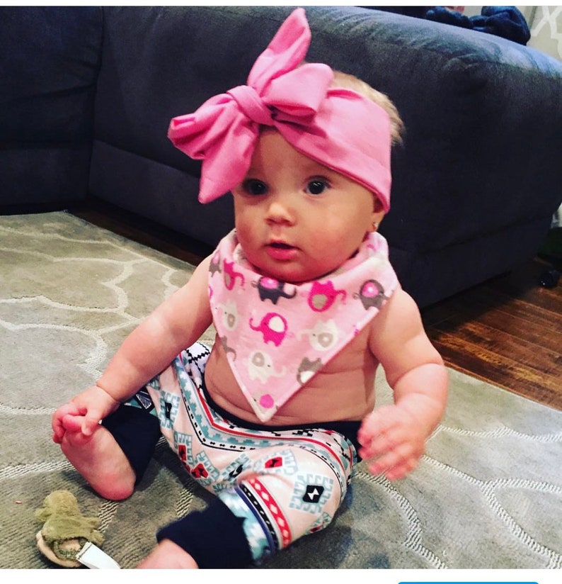May include: A baby wearing a pink headband with a bow, a pink and grey elephant print bib, and pink and blue patterned leggings.