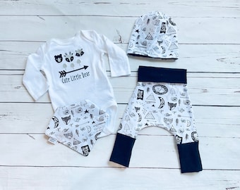 May include: A white baby outfit set with a black and white print featuring woodland animals, trees, and geometric shapes. The set includes a long-sleeved bodysuit with the text "Cute Little Bear", a pair of pants, a hat, and a bib.