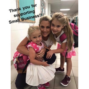May include: A woman and two young girls are smiling for the camera. The woman is wearing a white shirt and the girls are wearing pink and white dresses. The girls are holding backpacks. The text "Thank you for supporting Small Business!!" is superimposed on the image.