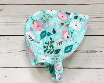 May include: A light blue sun hat with a floral print featuring pink and white flowers and green leaves. The hat has a pink button closure.