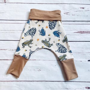 May include: White baby trousers with a brown waistband and brown cuffs. The trousers have a frog and bee print.