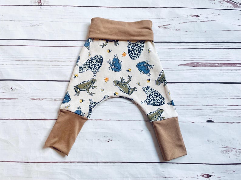 May include: White baby trousers with a brown waistband and brown cuffs. The trousers have a frog and bee print.