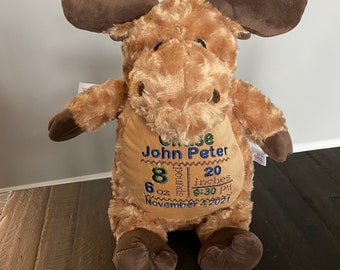 May include: A plush moose toy with tan fur and dark brown antlers and hooves. The belly features embroidered text: Chase John Peter, 8 pounds 6 oz, 20 inches, 9:30 PM, and November 4, 2021, stitched in blue and green thread on a tan patch.