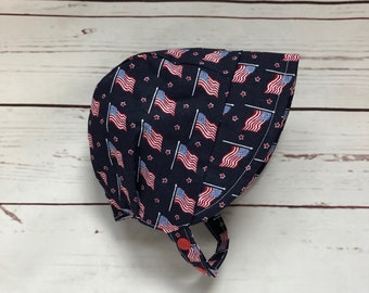 May include: A blue fabric baby bonnet with a white background and a pattern of red, white, and blue American flags. The bonnet has a red button on the strap.