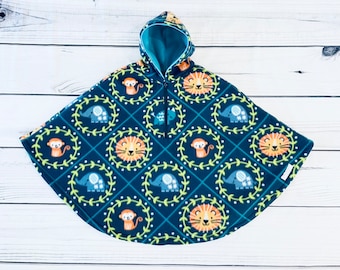 May include: A navy blue fleece poncho with a jungle animal design. The poncho features a repeating pattern of lions, elephants, and monkeys in circular wreaths. The hood and interior lining are teal, and it has a quarter-length zip.