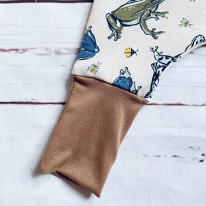 May include: A pair of brown and white trousers with a frog and bug print. The trousers have a brown cuff at the bottom.