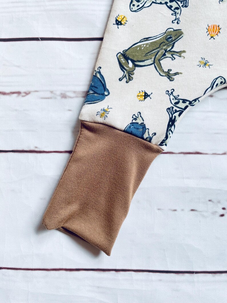 May include: A pair of brown and white trousers with a frog and bug print. The trousers have a brown cuff at the bottom.