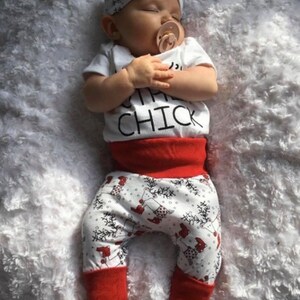 May include: A baby wearing a white onesie with the text "Little Chick" and red and white patterned trousers. The baby is wearing a white headband with a bow and has a pacifier in their mouth.