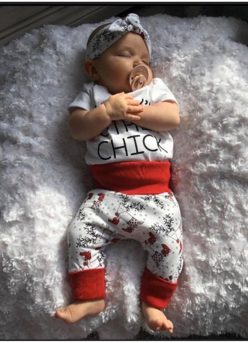May include: A baby wearing a white onesie with the text "Little Chick" and red and white patterned trousers. The baby is wearing a white headband with a bow and has a pacifier in their mouth.