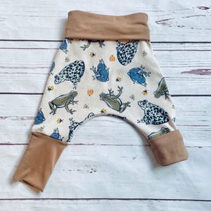 May include: White baby trousers with a brown waistband and a frog print. The frogs are green, blue and black with white spots.
