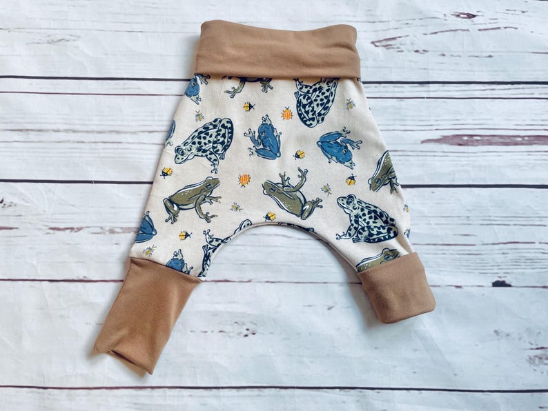 May include: White baby trousers with a brown waistband and a frog print. The frogs are green, blue and black with white spots.