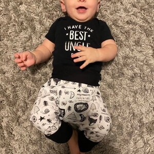 May include: A baby wearing a black short-sleeved onesie with the text "I have the best uncle" in white. The baby is also wearing black and white patterned trousers with a black waistband.