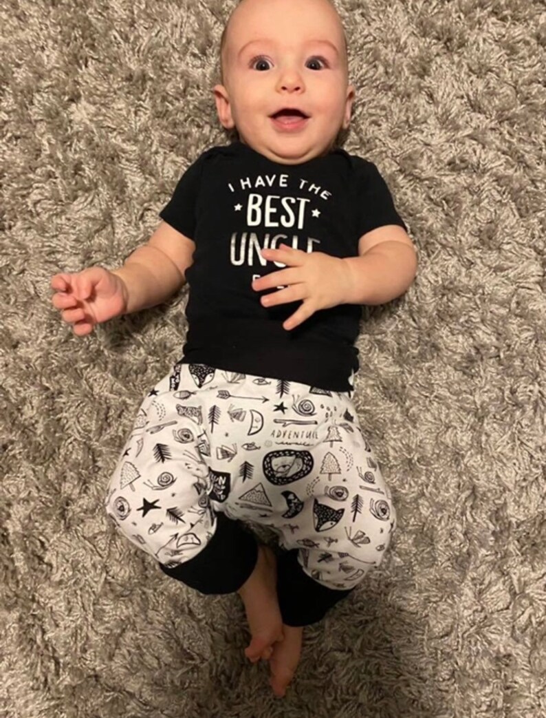 May include: A baby wearing a black short-sleeved onesie with the text "I have the best uncle" in white. The baby is also wearing black and white patterned trousers with a black waistband.