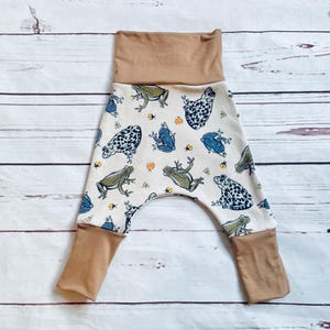May include: White baby trousers with a brown waistband and cuffs. The trousers have a pattern of frogs and insects.
