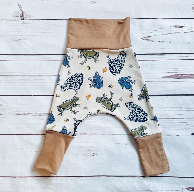 May include: White baby trousers with a brown waistband and cuffs. The trousers have a pattern of frogs and insects.