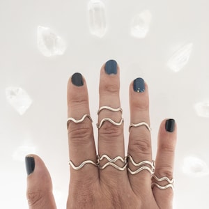 May include: A hand wearing several silver wave-shaped rings. The rings are stacked on each finger. The hand is painted with dark blue nail polish.