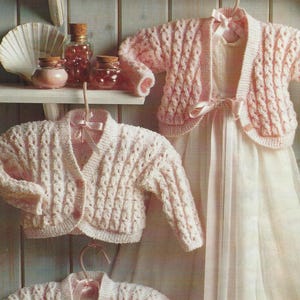 Puede incluir: Tres cárdigans de punto rosa para bebé con un patrón de trenza. Los cárdigans están colgados en perchas y están dispuestos en línea vertical.