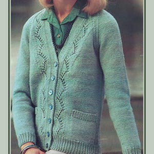 Può includere: Cardigan lavorato a maglia verde acqua con scollo a V, chiusura con bottoni e due tasche anteriori. Il cardigan presenta un motivo traforato decorativo sul davanti e sulle maniche. La persona indossa una camicia verde e una collana nera.