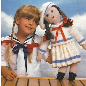 May include: A girl in a white sailor shirt with red and blue accents holds a knitted doll wearing a white sailor dress with blue and red accents. The doll has brown hair in pigtails and is wearing a white hat.