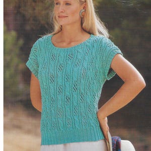 May include: A turquoise knitted top with a round neckline and short sleeves. The top features a textured, open-weave design. The woman is wearing a white skirt and holding a white hat with a blue and purple band.