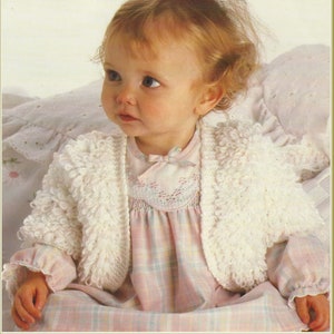 May include: A white crocheted baby cardigan with a ruffled edge. The cardigan is worn by a baby with blonde hair and is sitting on a white and pink blanket.