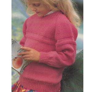 May include: A pink knitted sweater with ribbed detailing on the cuffs and neckline. The sweater has a horizontal ribbed design across the chest and upper sleeves. The person is holding a book with a picture of an egg.