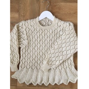 May include: A cream-coloured knitted jumper with a diamond pattern and scalloped edges. The jumper is hanging on a white hanger.