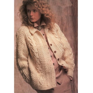 May include: A cream-colored cable knit cardigan with a button-up front and two pockets. The cardigan is worn over a white button-down shirt and brown pants.