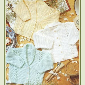May include: Three hand-knitted baby cardigans in pastel colors: pale yellow, white, and light blue. Each cardigan features a button-up front and intricate knit patterns. The garments are arranged on a wooden surface with decorative elements, suggesting a vintage or handmade aesthetic.