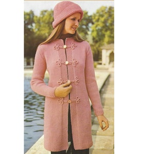 May include: A long pink knitted cardigan with a high collar and decorative knot buttons. The cardigan has long sleeves and a straight fit.